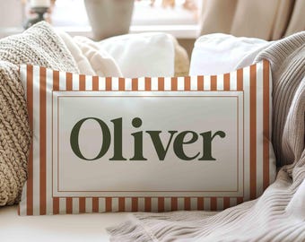 May include: A rectangular throw cushion featuring a white and burnt orange vertical striped pattern. The centre of the cushion displays a rectangular frame containing the name Oliver written in a dark green serif font on a light background.