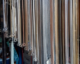 May include: A variety of silver, gold, and rose gold chains hanging from hooks. The chains are different lengths and thicknesses.