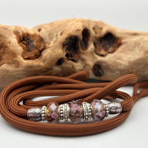 May include: A brown nylon cord necklace featuring three patterned glass beads interspersed with four silver-toned metal spacers. The beads display a swirling pink and white design, while the metal spacers have intricate engraved patterns.