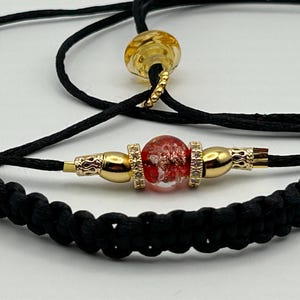 Op de afbeelding: Close-up van een zwart koordarmband met een rode en gouden kraal. De armband heeft goudkleurige metalen accenten en een gevlochten koordontwerp. Een tweede koord met een gele kraal is ook zichtbaar.