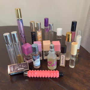 May include: A collection of various perfume bottles and fragrance vials of different shapes, sizes, and colors, including glass spray bottles, rollerballs, and a small rectangular box labeled Yes I Am, alongside a bright coral textured cylindrical tube.
