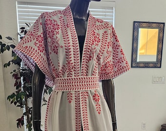 May include: White, floor-length kimono-style robe with red floral and leaf embroidery. The robe features a matching embroidered belt and trim along the edges. The garment is displayed on a mannequin, showcasing its flowing design and intricate details.