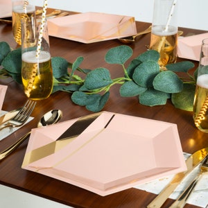 May include: A table setting with pink and gold geometric plates, gold cutlery, and champagne flutes with gold-striped straws. A green eucalyptus garland runs down the center of the dark wood table, creating an elegant and festive atmosphere.