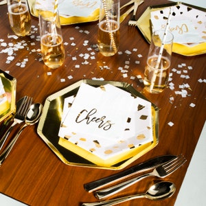 May include: Gold and white paper party plates with a gold confetti design and the word "Cheers" printed on the napkins. The plates are set on a wooden table with gold silverware and champagne flutes.