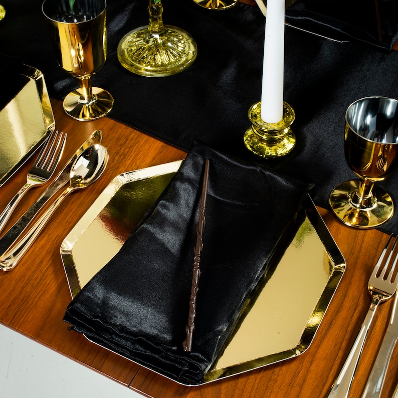 May include: A gold-rimmed, octagonal dinner plate with a black satin napkin and a black wand on top. The plate is set on a wooden table with a black tablecloth and gold silverware.