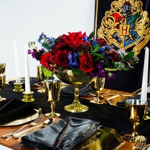 May include: A black table setting with gold goblets, candlesticks, and a gold bowl filled with red, purple, and blue flowers. The table is set with black napkins and gold chargers. A black and gold banner with the Hogwarts crest is in the background.