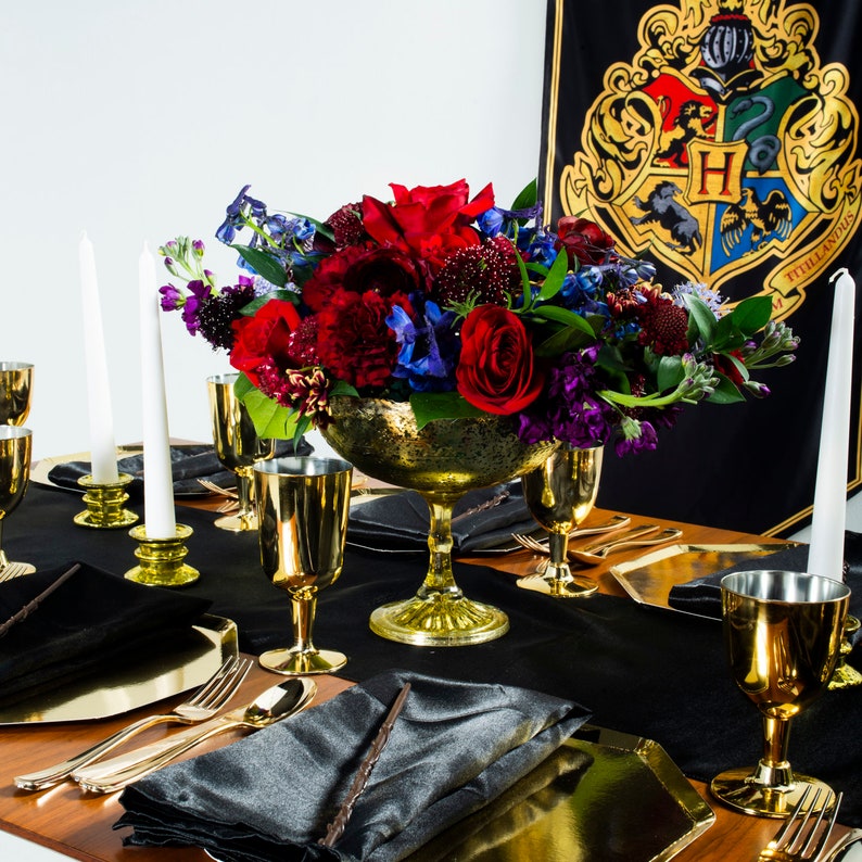 May include: A black table setting with gold goblets, candlesticks, and a gold bowl filled with red, purple, and blue flowers. The table is set with black napkins and gold chargers. A black and gold banner with the Hogwarts crest is in the background.