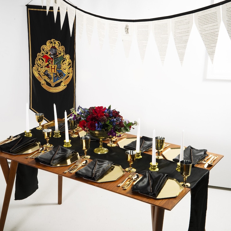 May include: A black and gold table setting with black napkins, gold chargers, and gold silverware. A centerpiece of red and purple flowers sits in the middle of the table. A black banner with the Hogwarts crest hangs on the wall behind the table.