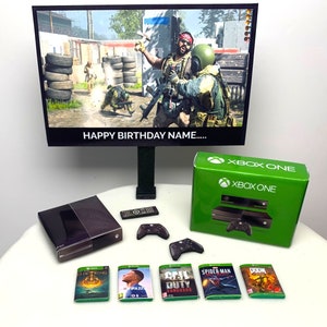 Peut inclure: Un ensemble cadeau d'anniversaire pour un fan de jeux vidéo. L'ensemble comprend une console Xbox One noire, une manette Xbox One noire, deux manettes Xbox One noires et vertes, un disque de jeu Xbox One noir et cinq étuis de jeux vidéo. Les étuis de jeux sont pour les jeux Elden Ring, Call of Duty: Vanguard, Spider-Man et Doom.