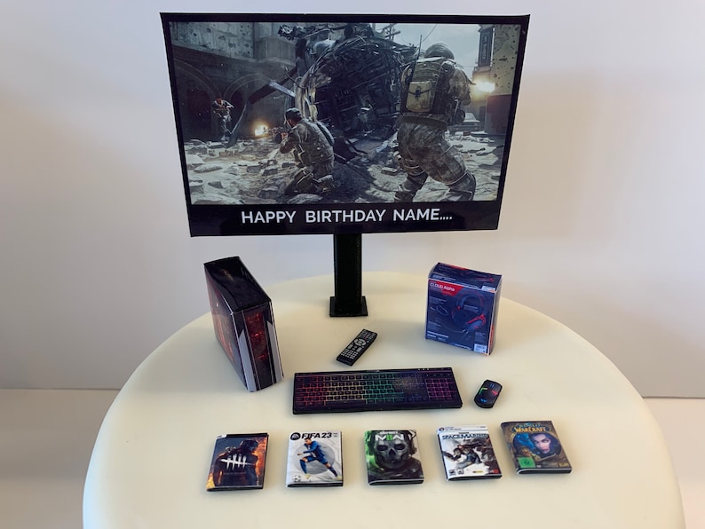 K&ouml;nnte beinhalten: Ein Miniatur-Gaming-Setup-Kuchentopper mit einem schwarzen Computermonitor, einer schwarzen Tastatur, einem schwarzen und roten Computergeh&auml;use, einer schwarzen Fernbedienung, einer schwarzen Maus, einer roten und schwarzen Headset-Box und sechs Spielverpackungen. Der Monitor zeigt eine Videospielszene mit Soldaten und einem Hubschrauber. Der Text "HAPPY BIRTHDAY NAME..." wird auf dem Monitor angezeigt.
