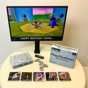 Può includere: Una torta di compleanno decorata con una console PlayStation, controller e cartucce di gioco. La torta è decorata con uno schermo che mostra un personaggio di videogioco. Il testo sullo schermo recita "HAPPY BIRTHDAY NAME..."