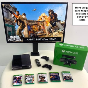 K&ouml;nnte beinhalten: Eine schwarze Xbox One Konsole mit einem gr&uuml;nen Controller und einem schwarz-gr&uuml;nen Spielgeh&auml;use. Die Konsole steht auf einem wei&szlig;en Kuchenst&auml;nder mit einem schwarz-gr&uuml;nen Xbox One Controller, einer schwarzen Fernbedienung und mehreren Spielgeh&auml;usen. Die Konsole steht vor einem schwarzen Computermonitor mit einem Bild des Videospiels Call of Duty: Black Ops Cold War auf dem Bildschirm. Der Monitor steht auf einem schwarzen St&auml;nder. Das Bild auf dem Bildschirm zeigt einen Soldaten, der eine Waffe h&auml;lt, und einen Hubschrauber, der am Himmel fliegt. Der Text "Happy Birthday Name..." ist auf dem Bildschirm zu sehen. Der Text "More unique cake toppers available in our ETSY store" befindet sich auf der rechten Seite des Bildes.