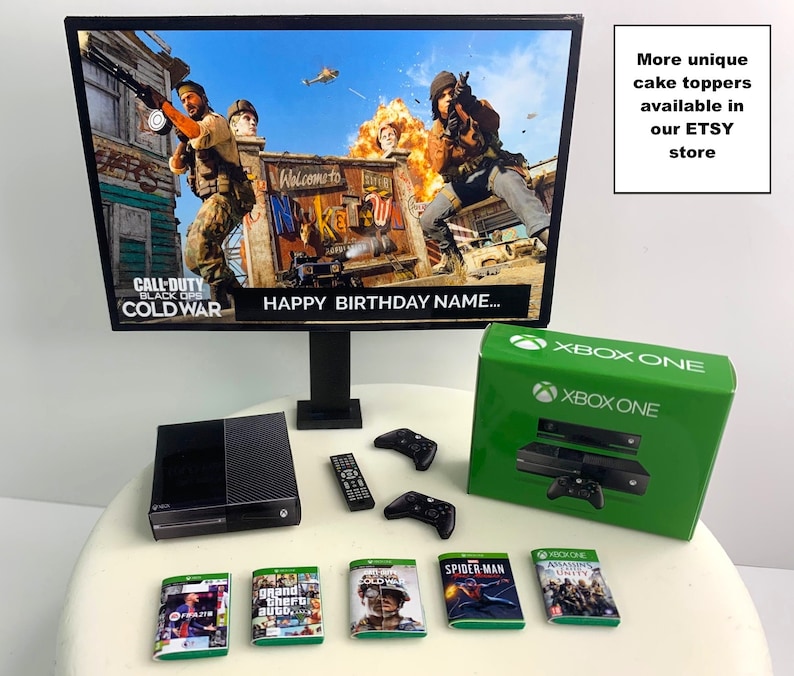 K&ouml;nnte beinhalten: Eine schwarze Xbox One Konsole mit einem gr&uuml;nen Controller und einem schwarz-gr&uuml;nen Spielgeh&auml;use. Die Konsole steht auf einem wei&szlig;en Kuchenst&auml;nder mit einem schwarz-gr&uuml;nen Xbox One Controller, einer schwarzen Fernbedienung und mehreren Spielgeh&auml;usen. Die Konsole steht vor einem schwarzen Computermonitor mit einem Bild des Videospiels Call of Duty: Black Ops Cold War auf dem Bildschirm. Der Monitor steht auf einem schwarzen St&auml;nder. Das Bild auf dem Bildschirm zeigt einen Soldaten, der eine Waffe h&auml;lt, und einen Hubschrauber, der am Himmel fliegt. Der Text "Happy Birthday Name..." ist auf dem Bildschirm zu sehen. Der Text "More unique cake toppers available in our ETSY store" befindet sich auf der rechten Seite des Bildes.