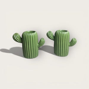 May include: Two green cactus-shaped ceramic planters. Each planter has a cylindrical body with vertical ridges and two arm-like protrusions. The planters are a light green color and have a hollowed-out top for planting.