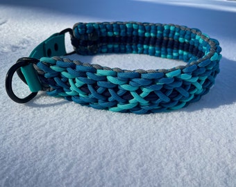 Puede incluir: Un collar de perro trenzado de color azul verdoso, azul y gris con un anillo en D de metal negro.