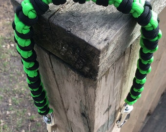 May include: A black and neon green paracord leash with a silver clip. The leash is coiled and resting on a wooden surface.