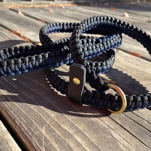 Op de afbeelding: Een zwarte en blauwe paracord hondenriem met een leren handvat en een gouden metalen ring. De riem is opgerold op een houten oppervlak.