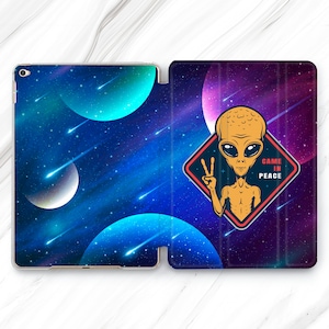 Puede incluir: Una funda para iPad con temática de galaxia azul y morada con un extraterrestre amarillo sosteniendo un signo de paz y una insignia roja y blanca que dice "Came in Peace".