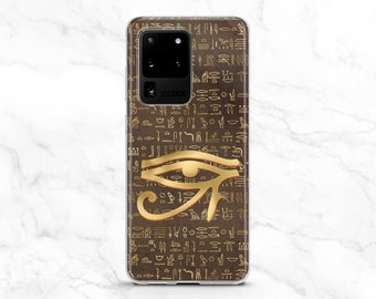 May include: A clear phone case with a brown background and gold hieroglyphics. A gold Eye of Horus symbol is in the center of the case.