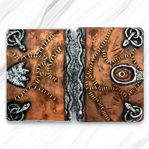 May include: A brown tablet case designed to look like a spell book. The case has a textured, stitched design with silver accents, including snakes and a central eye. The case is open, revealing the tablet screen.