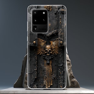 May include: A black phone case with a gold skull and wings design. The case has a textured surface and the text "100X" is visible on the back.