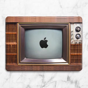 Könnte beinhalten: Ein Vintage-Fernseher mit Holzrahmen und einem schwarzen Apple-Logo auf dem Bildschirm. Das TV-Gerät hat ein Retro-Design mit Drehknöpfen und vertikalen Lamellen an den Seiten. Der Bildschirm ist hellgrau.