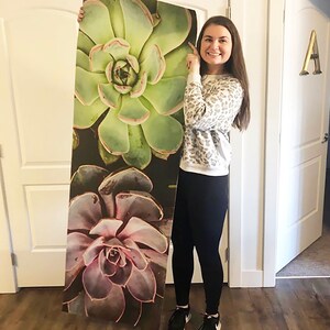 May include: A woman holds up a large printed yoga mat featuring a close-up of three succulent plants. The plants are green, purple, and brown with a dark background.