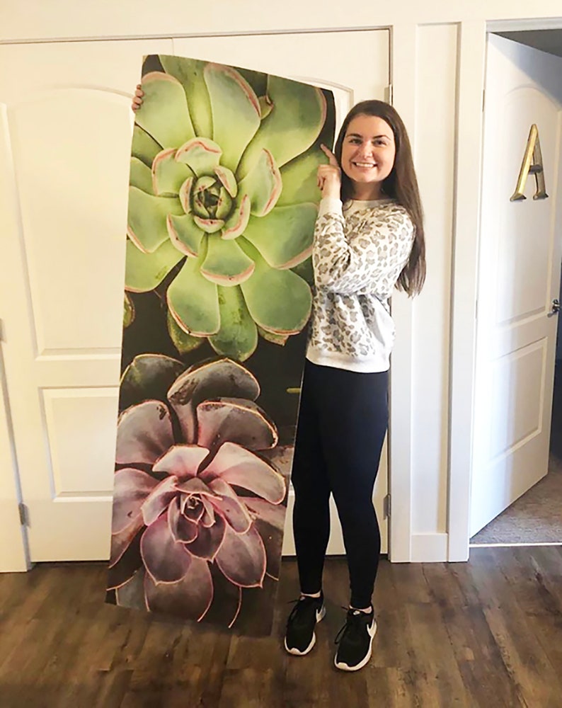 May include: A woman holds up a large printed yoga mat featuring a close-up of three succulent plants. The plants are green, purple, and brown with a dark background.