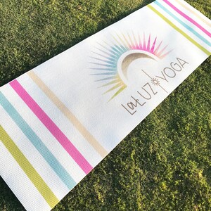 May include: A white yoga mat with colorful stripes and a sunburst design. The text "La Luz Yoga" is printed in brown on the mat.