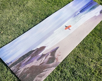 Puede incluir: Una esterilla de yoga rectangular con una escena de playa y un surfista. El diseño de la esterilla incluye una playa de arena, olas del océano y un surfista con una tabla de surf naranja. La esterilla está colocada sobre una superficie de hierba verde.