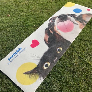 May include: A white yoga mat with a black cat and a brown and white dog peeking over the edge. The mat has colorful polka dots and the text "pumpkin. The best treat is a healthy life."