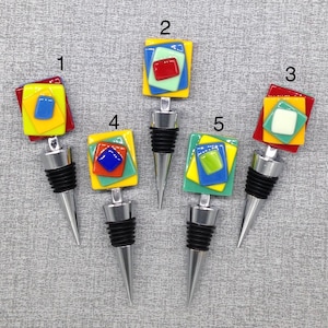 May include: Five colorful glass bottle stoppers with geometric designs in shades of red, yellow, blue, and green. Each stopper has a silver metal base.