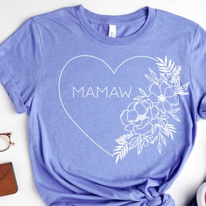 May include: A periwinkle blue t-shirt featuring a white heart design with the word "MAMAW" inside. The heart is outlined with a floral arrangement on the right side. The shirt is made of a soft material.