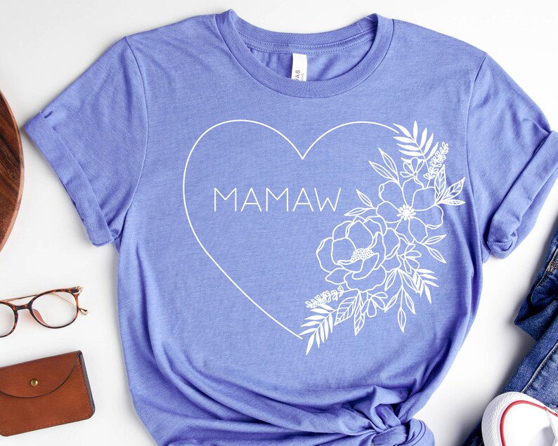 May include: A periwinkle blue t-shirt featuring a white heart design with the word "MAMAW" inside. The heart is outlined with a floral arrangement on the right side. The shirt is made of a soft material.