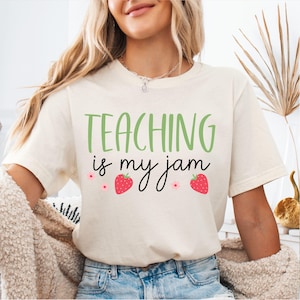May include: A cream-colored short-sleeve t-shirt featuring the green and black text "TEACHING is my jam" printed on the front. The design includes two red strawberries and two small pink flowers positioned below the text.