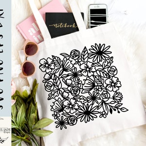 May include: A white tote bag with a black floral design. The design features a variety of flowers and leaves, all outlined in black. The bag is lying on a white surface.