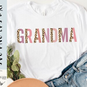 May include: A white t-shirt with the word "GRANDMA" printed in pink, peach, and brown leopard print letters.