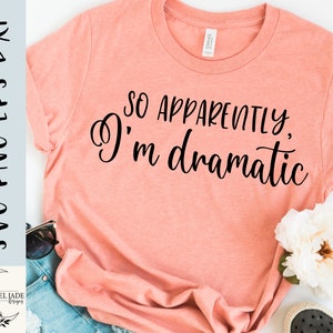 Może przedstawiać: Brzoskwiniowa koszulka z napisem "SO APPARENTLY, I'm dramatic" czcionką w kolorze czarnym. Koszulka wykonana jest z miękkiego materiału i jest elementem odzieży codziennej. Projekt to cyfrowy plik SVG-PNG-EPS-DXF.