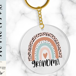 May include: A white circle keychain with a rainbow design and the word "Grandma" in black text. The rainbow has a pink, blue, and white color scheme with a black and white leopard print pattern.