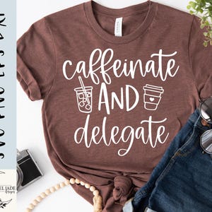 May include: A brown t-shirt with the words "caffeinate AND delegate" in white script. The design includes illustrations of an iced coffee and a to-go coffee cup. The shirt is paired with blue jeans and sunglasses.