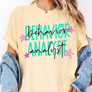 May include: A pale yellow short-sleeve t-shirt featuring the text "BEHAVIOR ANALYST" in large teal block letters with the words "behavior analyst" in black cursive script layered over them, accented by small pink stars with striped patterns.