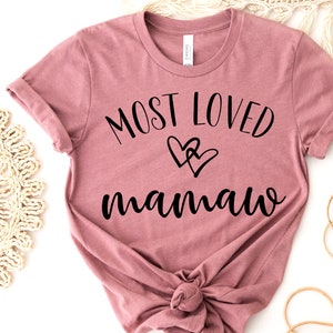 May include: Dusty rose t-shirt with rolled sleeves. The shirt features the text "MOST LOVED mamaw" in black script, with two overlapping heart graphics. The shirt is knotted at the bottom.
