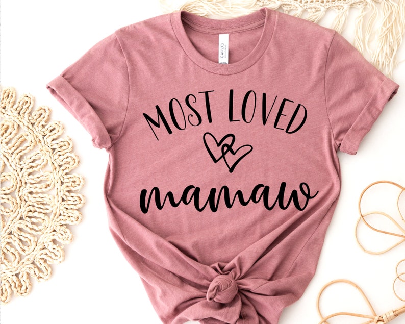 May include: Dusty rose t-shirt with rolled sleeves. The shirt features the text "MOST LOVED mamaw" in black script, with two overlapping heart graphics. The shirt is knotted at the bottom.
