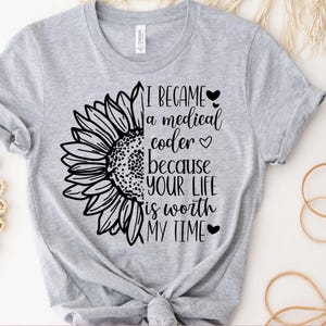 May include: A light gray t-shirt featuring a black and white sunflower design on the left side. The text "I BECAME a medical coder because YOUR LIFE is worth MY TIME" is printed in black. The words "INSTANT ACCESS" and "DIGITAL DOWNLOAD" are also visible.