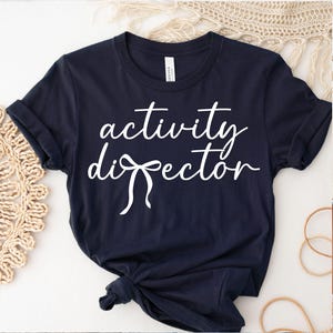 May include: Navy blue t-shirt with the words "activity director" in white script. The shirt has rolled-up sleeves and a knot at the bottom. The words "Instant Access" and "Digital Download" are at the top and bottom.