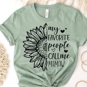 May include: Sage green t-shirt featuring a black sunflower graphic on the left side and the text "my favorite people call me Mamaw" on the right. The shirt is made of soft material.