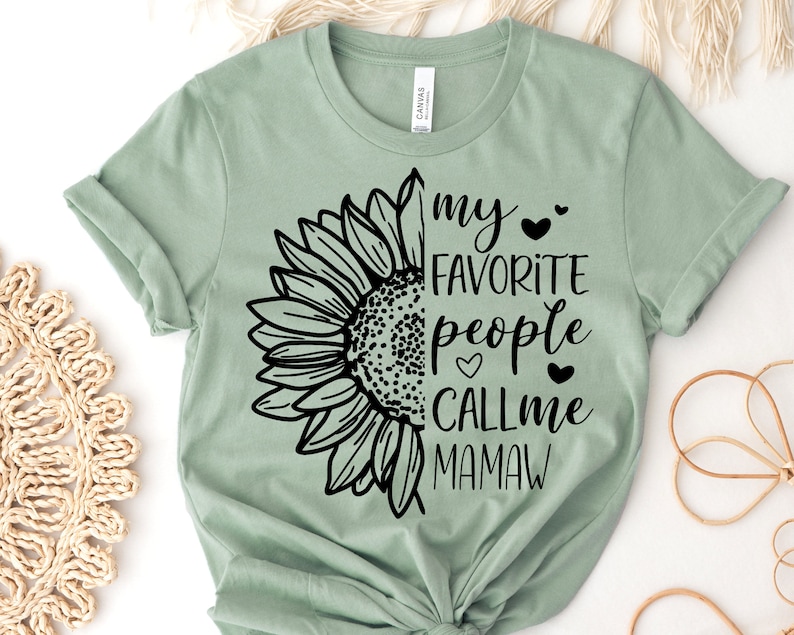 May include: Sage green t-shirt featuring a black sunflower graphic on the left side and the text "my favorite people call me Mamaw" on the right. The shirt is made of soft material.