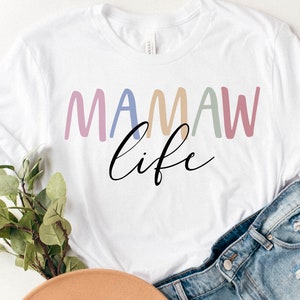 May include: White t-shirt with the text "MAMAW life" in colorful, rounded letters and a cursive font. The shirt is paired with denim shorts and a hat, suggesting a casual, everyday style. The design is simple and modern.