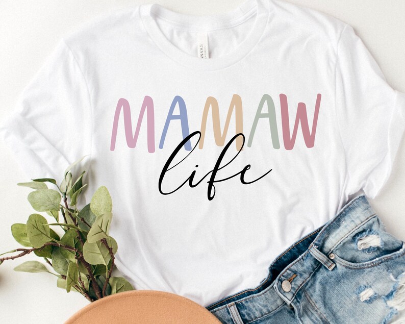 May include: White t-shirt with the text "MAMAW life" in colorful, rounded letters and a cursive font. The shirt is paired with denim shorts and a hat, suggesting a casual, everyday style. The design is simple and modern.