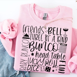 May include: A light pink sweatshirt featuring black text and graphics related to the game Bunco. The design includes the words friends, BELL, THREE OF A KIND, BUNCO, head table, LUCKY ROLL, DICE, and BABY BUNCO, along with icons of a bell, a drink, a scorecard, and dice.
